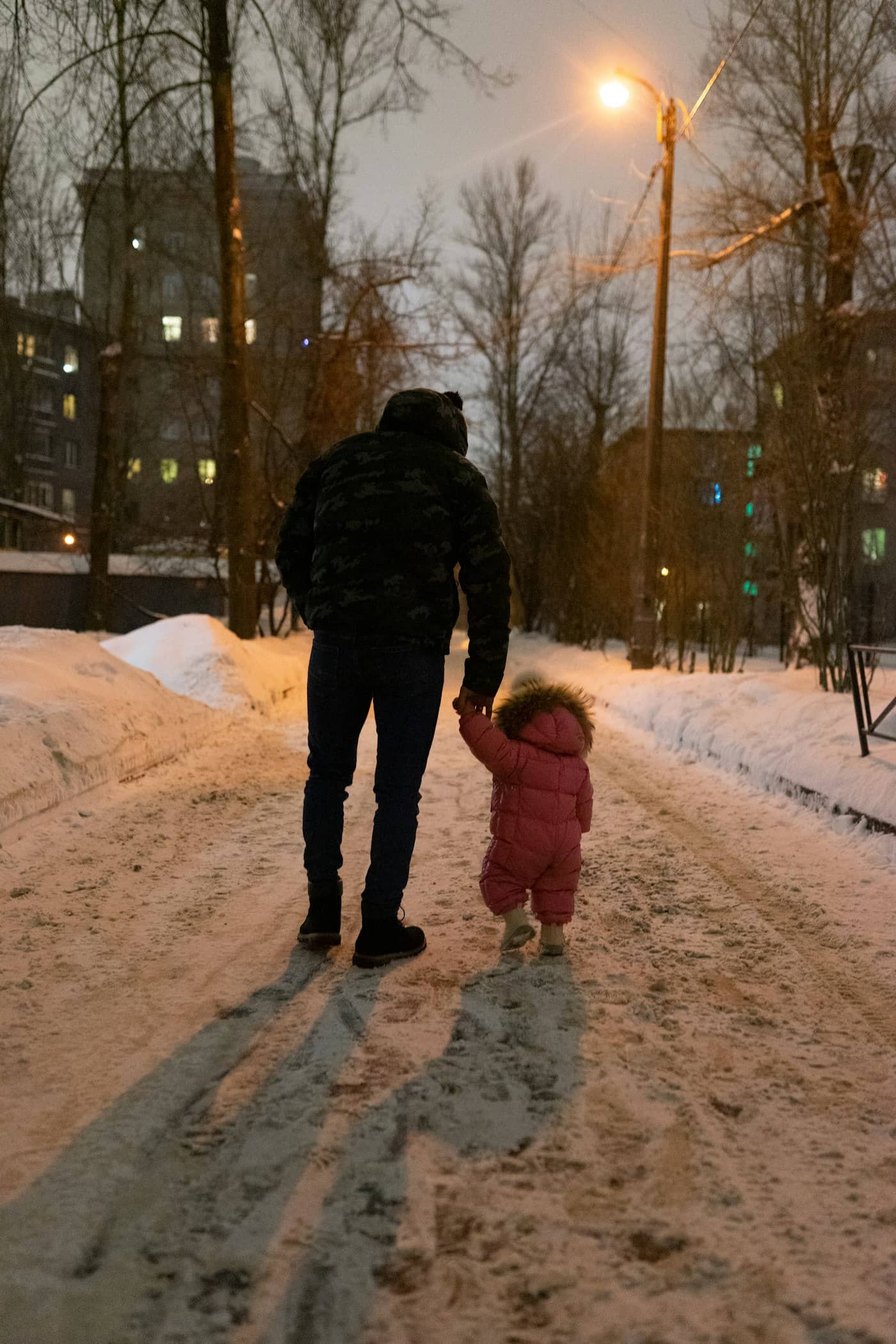 En forelder og et lite barn i vinterdress går hånd i hånd på en snødekt gate i skumringen